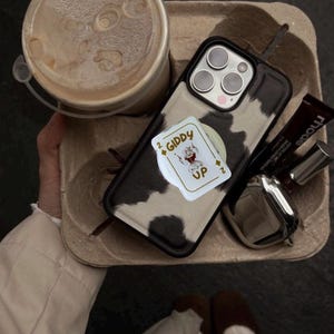 May include: A cow print phone case with a pop-up card holder featuring a playing card design that says "Giddy Up". The phone case is on a cardboard tray with a coffee drink, lipstick, and sunglasses.