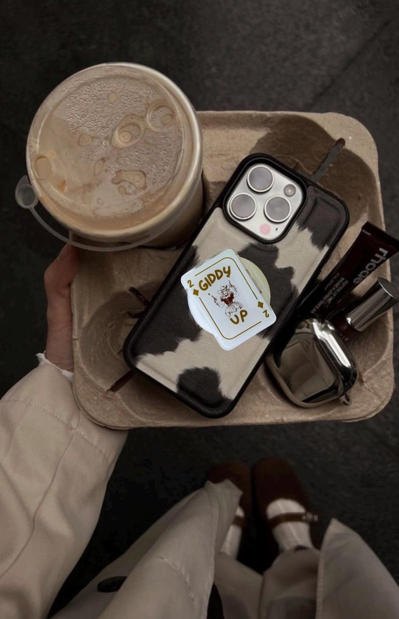 May include: A cow print phone case with a pop-up card holder featuring a playing card design that says "Giddy Up". The phone case is on a cardboard tray with a coffee drink, lipstick, and sunglasses.