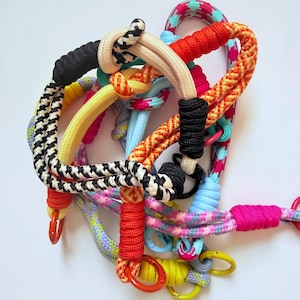 May include: A collection of multi-colored rope leashes featuring various braided patterns, including black and white houndstooth, solid orange, bright pink, and yellow. Each leash is secured with tightly wrapped cord accents and metal rings.