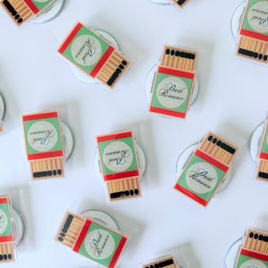 May include: An overhead shot shows multiple matchbox-shaped magnets. Each magnet features a red and green design with the words "Dark Romance" in a circular emblem. The matches are depicted with brown sticks and black tips.