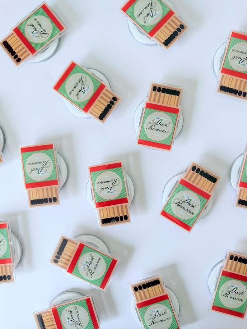May include: An overhead shot shows multiple matchbox-shaped magnets. Each magnet features a red and green design with the words "Dark Romance" in a circular emblem. The matches are depicted with brown sticks and black tips.