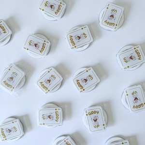 May include: White phone grips with a playing card design featuring a cartoon cowboy and the words "GIDDY UP". The grips are round and have a pop-out feature for easy handling. The card design includes the number "2" and diamond symbols.