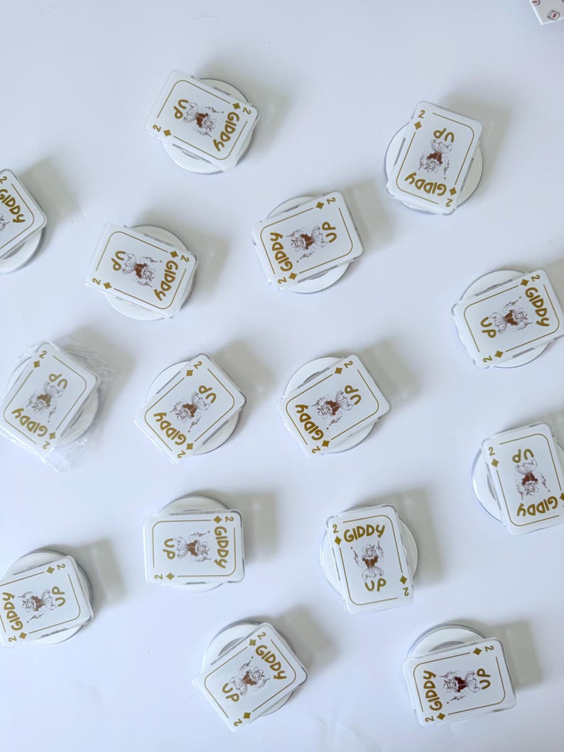 May include: White phone grips with a playing card design featuring a cartoon cowboy and the words "GIDDY UP". The grips are round and have a pop-out feature for easy handling. The card design includes the number "2" and diamond symbols.