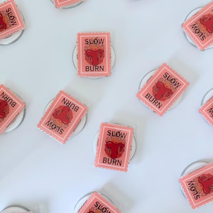 May include: A collection of pink phone grips with a rectangular design. Each grip features a red heart with flames and the words "SLOW BURN" and "BURN" in black text. The grips are arranged on a white surface.