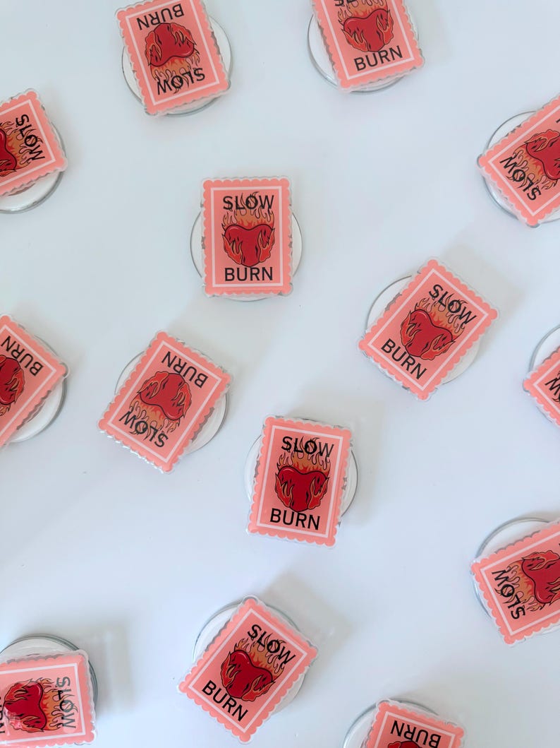 May include: A collection of pink phone grips with a rectangular design. Each grip features a red heart with flames and the words "SLOW BURN" and "BURN" in black text. The grips are arranged on a white surface.
