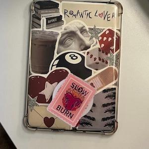 May include: Clear phone case decorated with various stickers. Stickers include a stack of books, a coffee cup, a bust, an eight ball, a heart, a stamp with the words "SLOW BURN", and a pair of eyes. The words "Romantic Lover" are also visible.