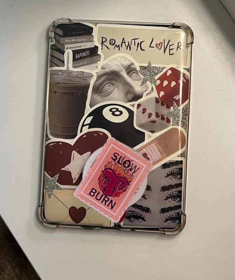 May include: Clear phone case decorated with various stickers. Stickers include a stack of books, a coffee cup, a bust, an eight ball, a heart, a stamp with the words "SLOW BURN", and a pair of eyes. The words "Romantic Lover" are also visible.