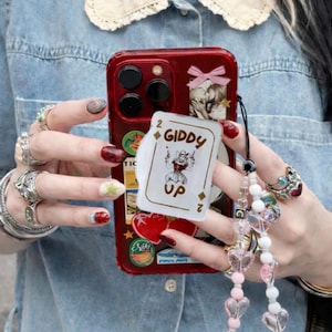 May include: Red phone case adorned with stickers and a playing card that reads "Giddy Up". The case is held by a person wearing multiple rings and a beaded phone charm with heart-shaped beads. The person is wearing a denim shirt.