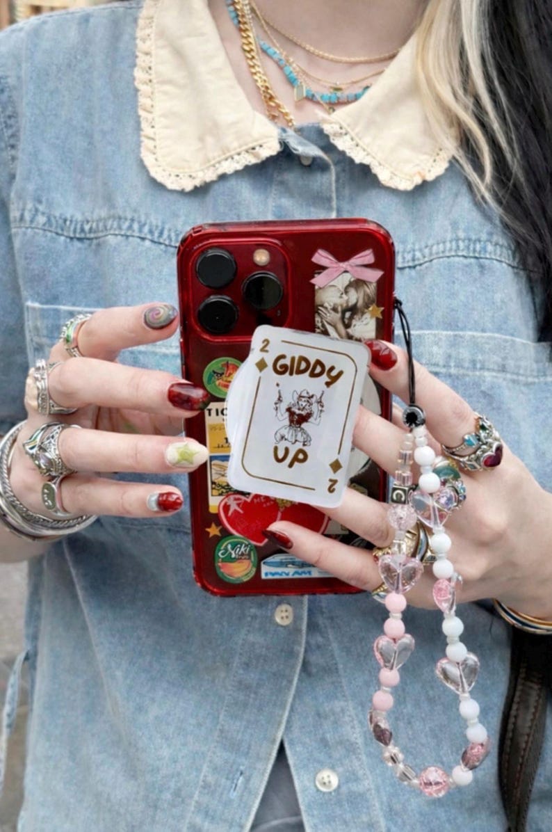May include: Red phone case adorned with stickers and a playing card that reads "Giddy Up". The case is held by a person wearing multiple rings and a beaded phone charm with heart-shaped beads. The person is wearing a denim shirt.