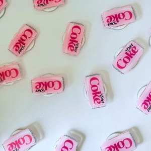 May include: Pink Diet Coke can-shaped phone grips with the Diet Coke logo in white. The grips are on a white surface. The grips have a silver glitter border.