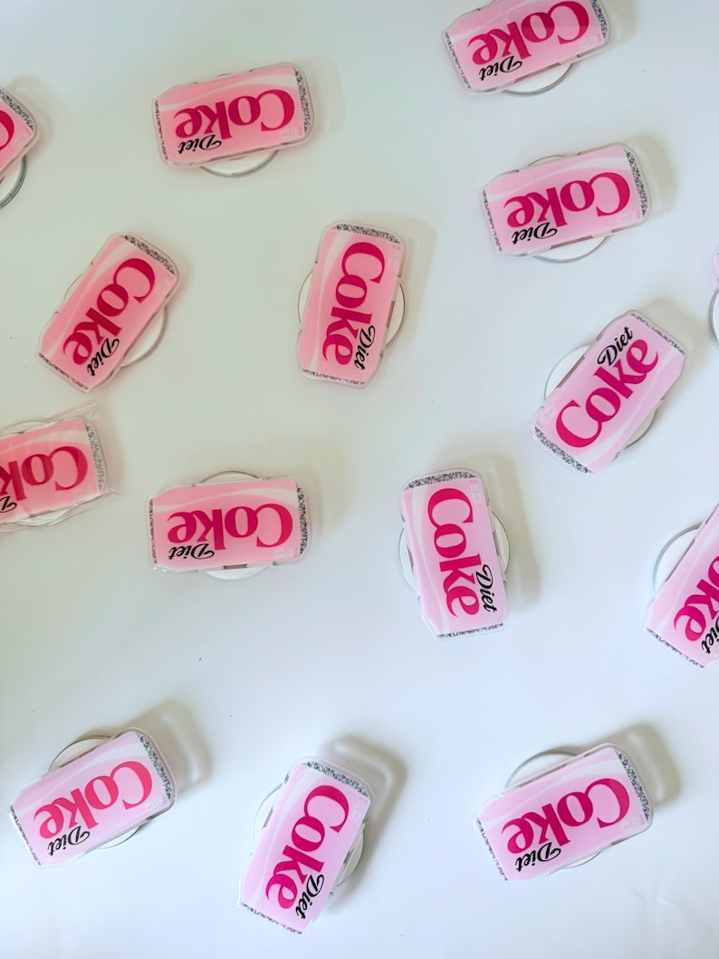 May include: Pink Diet Coke can-shaped phone grips with the Diet Coke logo in white. The grips are on a white surface. The grips have a silver glitter border.