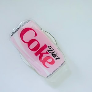May include: A Diet Coke phone grip with a pink and white design. The words "Coke" and "Diet" are printed in red and black, respectively. The grip has a clear, glittery border.