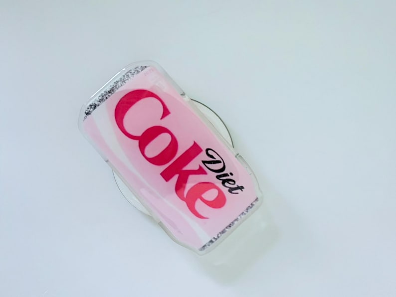 May include: A Diet Coke phone grip with a pink and white design. The words "Coke" and "Diet" are printed in red and black, respectively. The grip has a clear, glittery border.