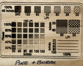 May include: A laser engraving test chart on a wood surface. The chart shows the effects of different power settings, feed rates, and line per inch (LPI) settings on the engraving quality. The chart is labeled "PINE + BORAX".