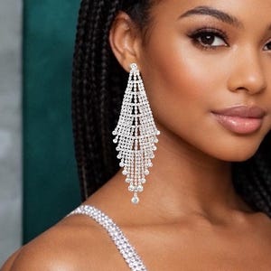 Peut inclure: Une femme aux longues tresses noires porte une robe argentée ornée de cristaux et des boucles d'oreilles pendantes. Les boucles d'oreilles sont une cascade de petits cristaux, et la robe a un décolleté plongeant. L'arrière-plan est un mélange de turquoise et de gris.