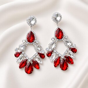 May include: A pair of chandelier earrings featuring a circular top stud, a central pear-shaped red crystal, and a dangling frame adorned with alternating clear and red teardrop-shaped crystals set in a silver-toned metal base.