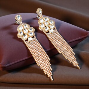 May include: A pair of gold-coloured, dangling earrings. Each earring has a cluster of clear, oval-shaped stones at the top, with a cascade of smaller, sparkling stones hanging below. The earrings are set against a dark purple cushion and a brown background.