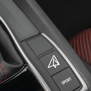 May include: A close-up of a car's interior showing a black button with a white airplane icon and the word "SPORT" on a black button below it.