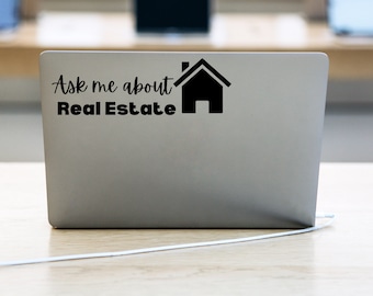 May include: A silver laptop with a black sticker that reads "Ask me about Real Estate" in a script font, accompanied by a house icon. The text is centered, with the house icon to the right.