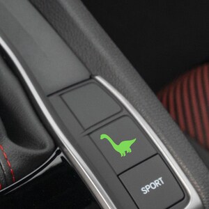 May include: A black car interior with a green dinosaur graphic on a button labelled "SPORT".