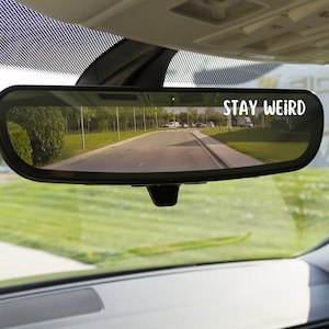 May include: A car's rearview mirror with the words "STAY WEIRD" in white. The mirror reflects a road with green grass and trees on either side. The car's interior is visible.
