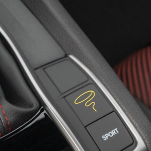 May include: Close-up of a car's interior showing a black button with a yellow icon of a circle with a squiggly line inside. The button is labeled "SPORT".