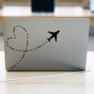 Może przedstawiać: Srebrny laptop z czarną naklejką samolotu lecącego w kropkowanej linii, która tworzy kształt serca.