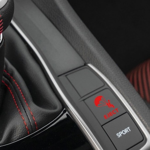 Op de afbeelding: Een zwart auto-interieur met een rode gestikte versnellingspook en twee knoppen. De knop links heeft een rood parachute-icoon en de tekst "EJECT". De knop rechts heeft de tekst "SPORT".