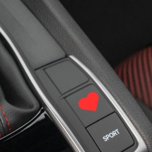Könnte beinhalten: Nahaufnahme des Innenraums eines Autos mit einem roten herzförmigen Knopf mit der Aufschrift "SPORT".