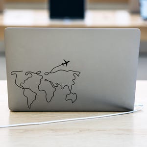 Könnte beinhalten: Ein silberner Laptop mit einem schwarzen Weltkarten- und Flugzeugaufkleber auf dem Deckel.