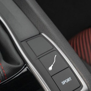 May include: A black car interior with a silver trim and a black button with a white broom icon. The button is labeled "SPORT".
