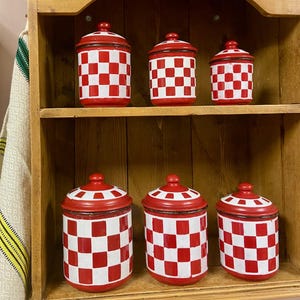Op de afbeelding: Een set van zes rood-witte geblokte keukencanners met bijpassende deksels. De canisters staan op een houten plank, wat een klassieke, vintage uitstraling geeft. De deksels hebben een rode knop.