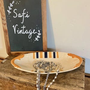 Peut inclure: Plat ovale en céramique crème avec des rayures orange et bleu marine, contenant un collier de perles. Le plat est posé sur une caisse en bois, avec un tableau noir indiquant "Safi Vintage".