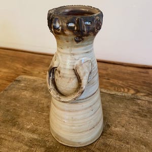 Può includere: Un vaso in ceramica fatto a mano con un design a forma di volto. Il vaso è beige con accenti marroni e una superficie strutturata. Presenta un volto con occhi, naso e bocca nella parte superiore. Un manico è attaccato al lato. Il vaso è su una superficie di legno.
