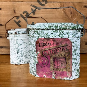 Op de afbeelding: Twee vintage lunchboxen van groen en wit emaille met metalen handvatten. De grotere doos heeft een roze en wit etiket met de tekst "L'ideal c'est la Arrival Pratique Unique Moderne".