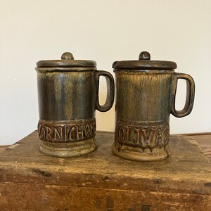 May include: Two cylindrical ceramic jars with lids and side handles, featuring a dark brown and tan drip glaze. The left jar has the word CORNICHONS embossed on the front, and the right jar has the word OLIVES embossed on the front.