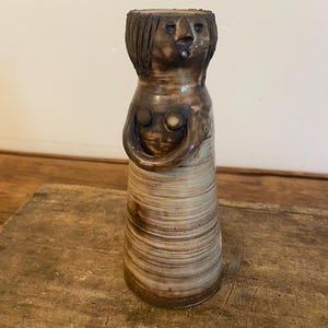 Può includere: Un vaso in ceramica con un volto e un corpo umanoide. Il vaso ha una combinazione di colori marrone e beige, con una superficie strutturata. La testa presenta un volto e capelli dettagliati, mentre il corpo ha una forma arrotondata con due dettagli circolari.
