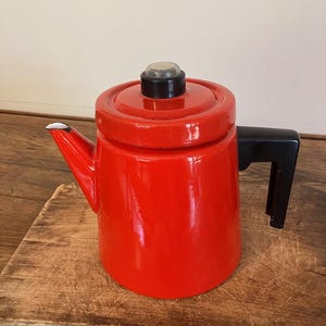 Puede incluir: Una cafetera de esmalte rojo brillante con acabado lustroso, asa negra y tapa roja a juego rematada con un pomo circular transparente. El pico vertedor también es rojo y presenta un pequeño borde blanco en la punta del mismo.