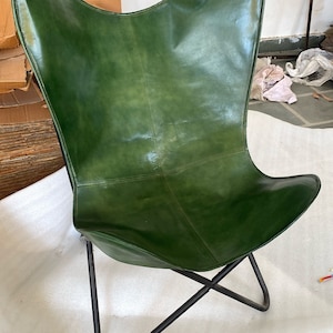 May include: A green leather butterfly chair with a black metal frame. The chair is folded and sitting on a white surface.