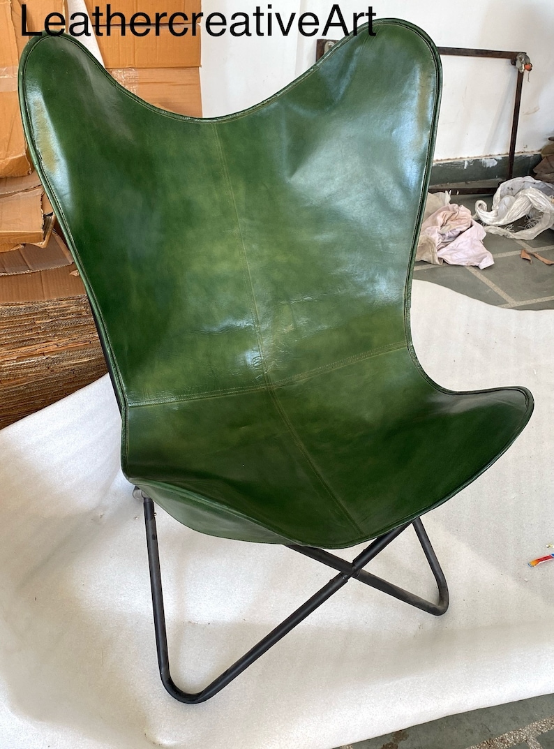 May include: A green leather butterfly chair with a black metal frame. The chair is folded and sitting on a white surface.