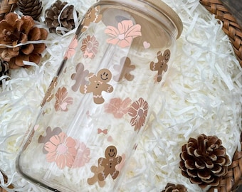 May include: A clear glass tumbler with a bamboo lid and straw, decorated with gingerbread men and floral designs in shades of brown and pink. The tumbler is set in a woven basket with white paper filler and pinecones.