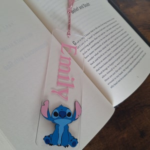 May include: A clear acrylic bookmark with a pink tassel. The bookmark has the name "Emily" written in pink lettering and a blue Stitch character from the Disney movie Lilo & Stitch.