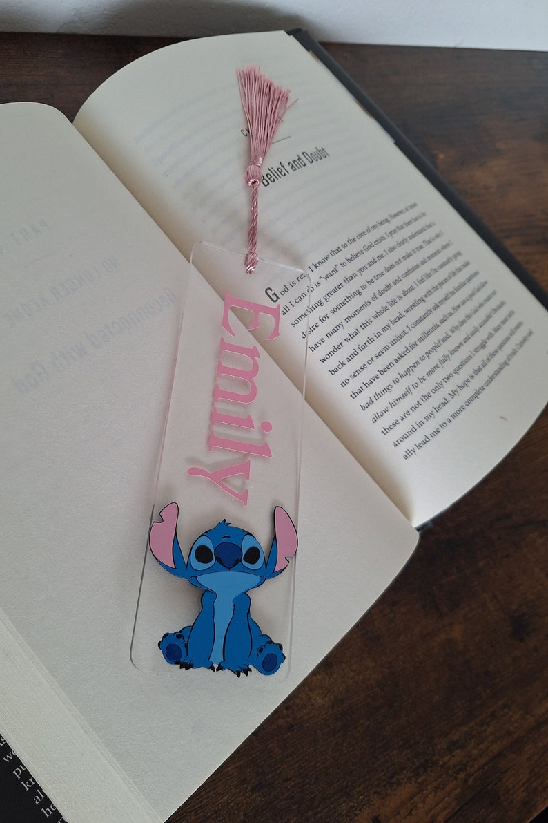 May include: A clear acrylic bookmark with a pink tassel. The bookmark has the name "Emily" written in pink lettering and a blue Stitch character from the Disney movie Lilo & Stitch.