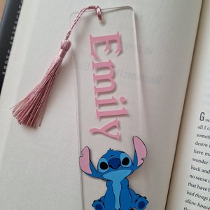 May include: Pink acrylic bookmark with the name "Emily" in pink lettering and a blue Stitch character from Lilo & Stitch. The bookmark has a pink tassel.