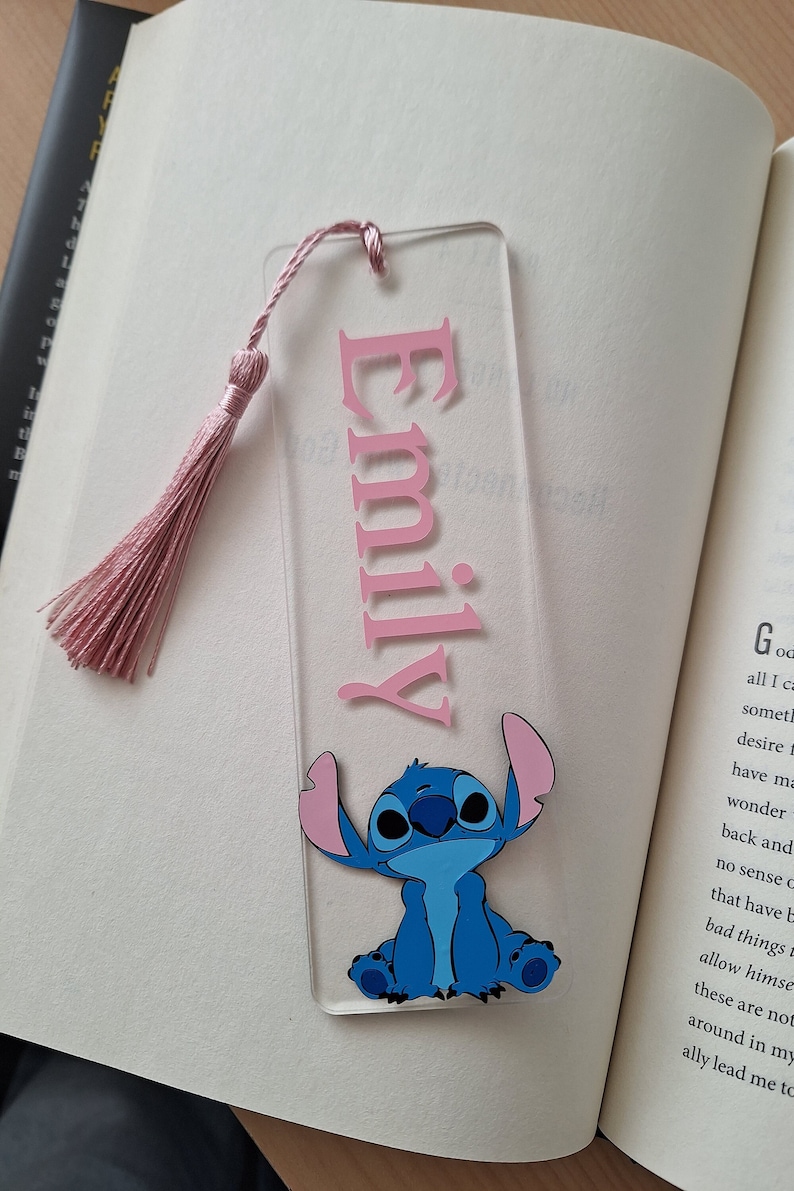 May include: Pink acrylic bookmark with the name "Emily" in pink lettering and a blue Stitch character from Lilo & Stitch. The bookmark has a pink tassel.