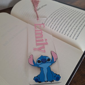 May include: A clear acrylic bookmark with a pink tassel. The bookmark features the name "Emily" in pink lettering and a blue cartoon character with pink ears.