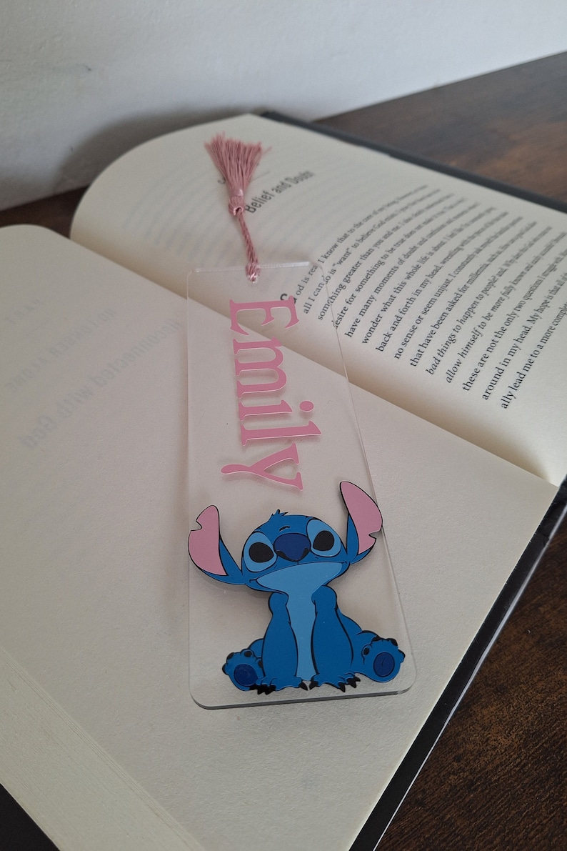 May include: A clear acrylic bookmark with a pink tassel. The bookmark features the name "Emily" in pink lettering and a blue cartoon character with pink ears.