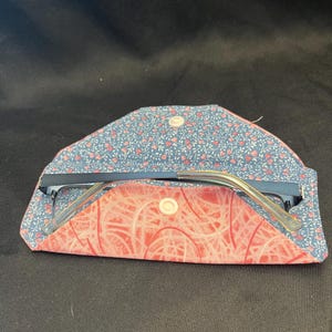 May include: A fabric glasses case with a blue floral exterior and a coral-colored interior featuring a white abstract line pattern. The case is open, revealing a pair of dark-framed glasses resting inside, and is secured by a white plastic snap button.