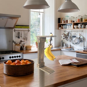 Puede incluir: Un colgador de plátanos de madera con un acabado desgastado. El colgador está montado en una encimera y tiene un gancho para colgar plátanos. Un racimo de plátanos cuelga del gancho.