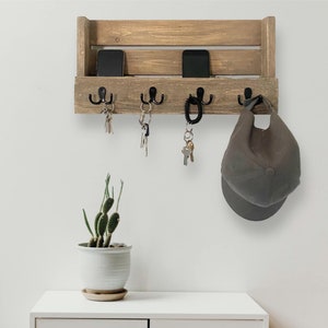 Könnte beinhalten: Rustikaler, an der Wand montierter Schlüsselhalter aus Holz mit einem Regal darüber zur Aufbewahrung. Der Schlüsselhalter hat vier Haken und das Regal hat zwei Schlitze für Telefone oder andere kleine Gegenstände. Der Schlüsselhalter ist in einem hellen Braunton gebeizt und hat eine Used-Look-Optik.